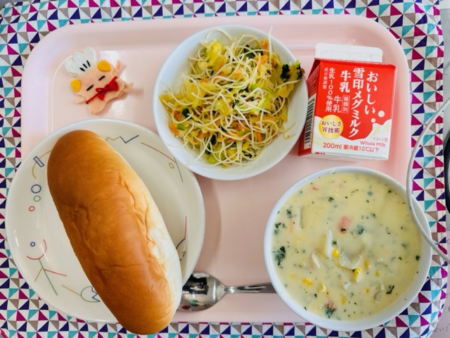 ピンクのトレーに給食が右上から反時計回りで牛乳、ビーフンソテー、コック帽をかぶった布製マスコット、あじつけパン、コーンシチューと並んでいる写真