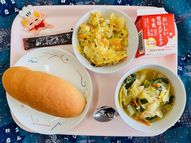 ピンクのトレーに給食が右上から反時計回りで牛乳、じゃがいものチーズ焼き、コック帽をかぶった布製マスコット、チョコレートパテ、コッペパン、肉団子とキャベツのスープと並んでいる写真