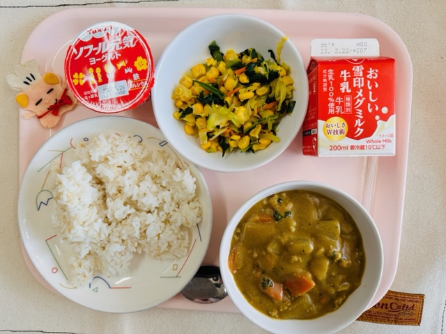 ピンクのトレーに給食が右上から反時計回りで牛乳、やさいソテー、ヨーグルト、コック帽をかぶった布製マスコット、ごはん、カレーのルーが並んでいる写真