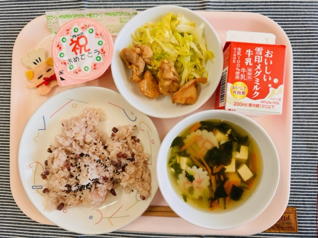ピンクのトレーに給食が右上から反時計回りで牛乳、キャベツソテー、とりにくのからあげ、デザート、コック帽をかぶった布製マスコット、せきはん（ゴマ塩）、すまし汁と並んでいる写真