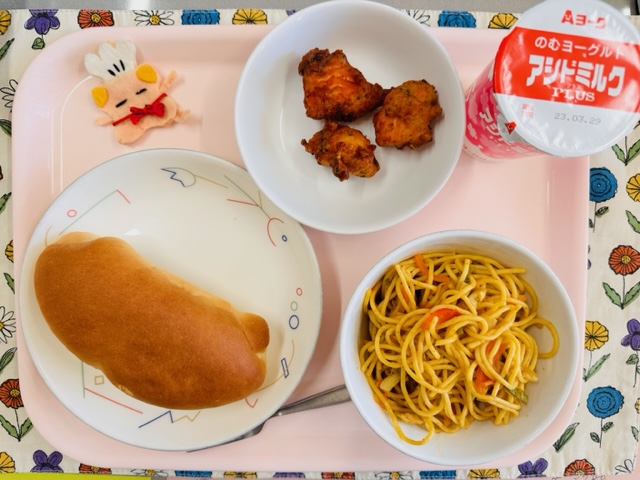 ピンクのトレーに給食が右上から反時計回りで牛乳、鮭のふわっと揚げ、コック帽をかぶった布製マスコット、ソフトフランスパン、カレー風味スパゲッティと並んでいる写真