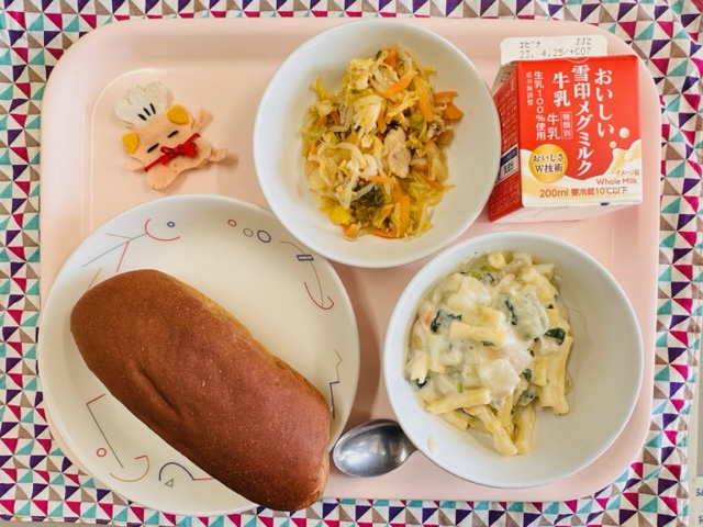 ピンクのトレーに給食が右上から反時計回りで牛乳、はるキャベツのサラダ、コック帽をかぶった布製マスコット、くろパン、マカロニのクリーム煮と並べられている写真