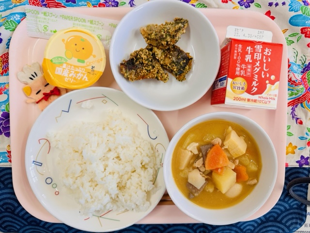 ピンクのトレーに給食が右上から反時計回りで牛乳、あじのごまあげ、国産みかんゼリー、コック帽をかぶった布製マスコット、ごはん、はるやさいのうまにと並べられている写真