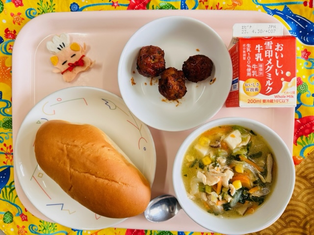 ピンクのトレーに給食が右上から反時計回りで牛乳、ビーンズミートボール、コック帽をかぶった布製マスコット、ミルクパン、コーン中華スープと並べられている写真