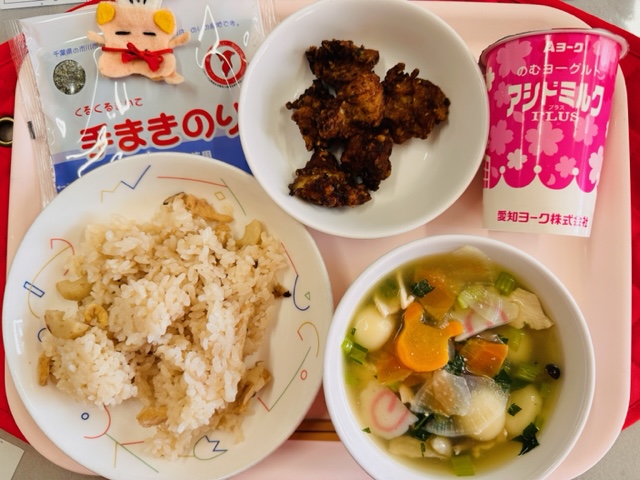 ピンクのトレーに給食が右上から反時計回りでのむヨーグルト、ぶたにくのふうみあげ、コック帽をかぶった布製マスコット、手まきのり、さくらずし、しらたまじると並べられている写真