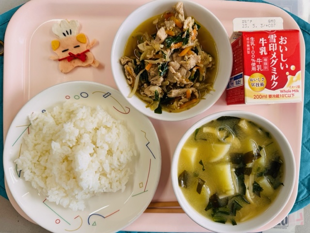 ピンクのトレーに給食が右上から反時計回りで牛乳、野菜入り焼肉、コック帽をかぶった布製マスコット、ごはん、みそしるの具と並べられている写真