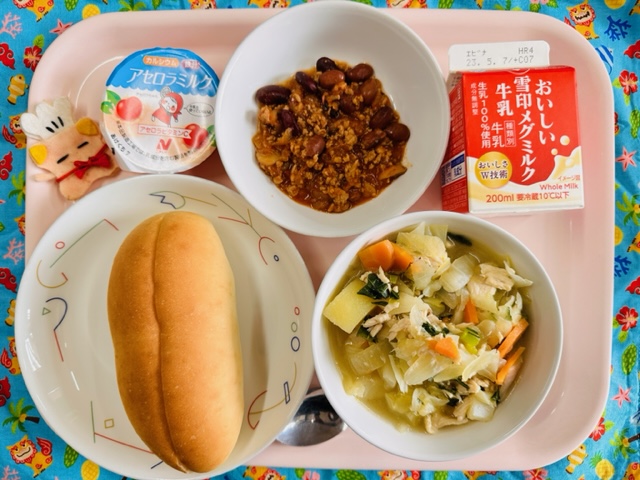 ピンクのトレーに給食が右上から反時計回りで牛乳、チリコンカン、アセロラミルク、コック帽をかぶった布製マスコット、パン、やさいのスープ煮と並べられている写真