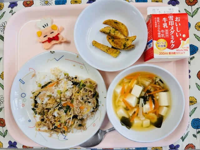 ピンクのトレーに給食が右上から反時計回りで牛乳、フライドポテト、コック帽をかぶった布製マスコット、ビビンパ、わかめスープと並べられている写真