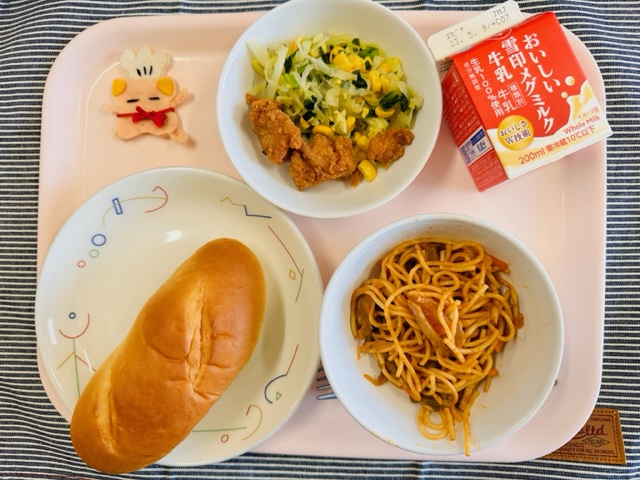 ピンクのトレーに給食が右上から反時計回りで牛乳、とりにくのからあげとミックスソテー、コック帽をかぶった布製マスコット、ミルクパン、スパゲッティナポリタンと並べられている写真