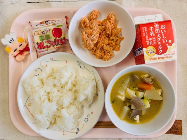 ピンクのトレーに給食が右上から反時計回りで牛乳、さけそぼろ、りんごのタルト、コック帽をかぶった布製マスコット、ごはん、とんじると並べられている写真