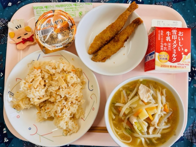 ピンクのトレーに給食が右上から反時計回りで牛乳、ししゃもフライ、みかんゼリー、コック帽をかぶった布製マスコット、たけのこごはん、あんかけじると並べられている写真