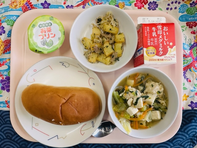 ピンクのトレーに給食が右上から反時計回りで牛乳、ジャーマンポテト、お茶プリン、コック帽をかぶった布製マスコット、あじつけパン、キャベツとにくだんごのスープと並べられている写真