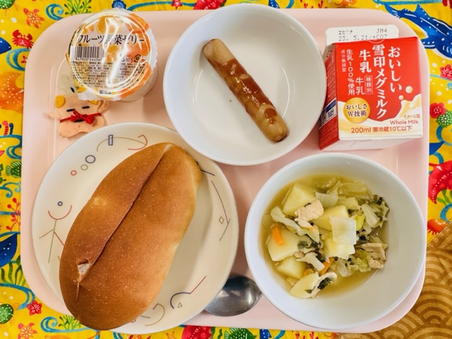 ピンクのトレーに給食が右上から反時計回りで牛乳、ウインナー、フルーツ野菜ゼリー、コック帽をかぶった布製マスコット、パン、やさいのスープ煮と並べられている写真