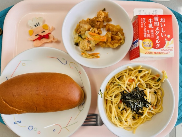 ピンクのトレーに給食が右上から反時計回りで牛乳、とりにくのマリネ、コック帽をかぶった布製マスコット、くろパン、わふうスパゲッティと並べられている写真