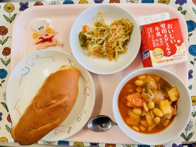 ピンクのトレーに給食が右上から反時計回りで牛乳、きりぼしだいこんのサラダ、コック帽をかぶった布製マスコット、ミルクパン、ポークビーンズと並べられている写真