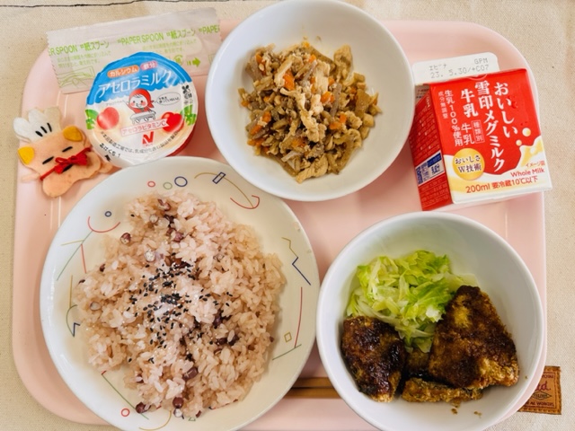 ピンクのトレーに給食が右上から反時計回りで牛乳、とりきんぴらごぼう、アセロラミルク、コック帽をかぶった布製マスコット、せきはんとごましお、あじフライと並べられている写真