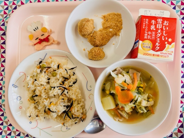 ピンクのトレーに給食が右上から反時計回りで牛乳、きなこドーナツ、コック帽をかぶった布製マスコット、ひじきピラフ、やさいスープと並べられている写真