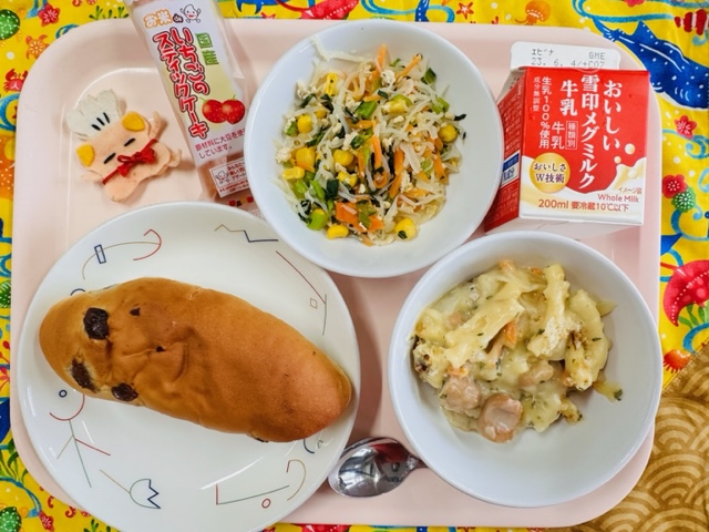 ピンクのトレーに給食が右上から反時計回りで牛乳、しらたきソテー、いちごのスティックケーキ、コック帽をかぶった布製マスコット、ぶどうパン、ウインナービーンズグラタンと並べられている写真