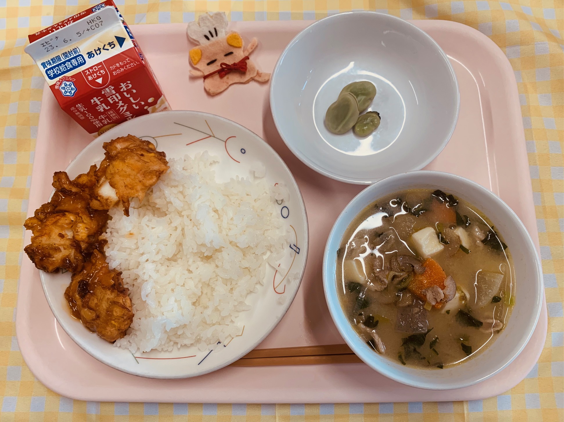 ピンクのトレーに給食が右上から反時計回りでそらまめ、コック帽をかぶった布製マスコット、牛乳、かきあげどん、あっさりとんじると並べられている写真