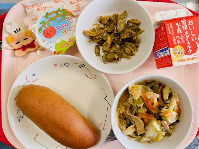 ピンクのトレーに給食が右上から反時計回りで牛乳、ごぼうのあまからだれからめ、りんごゼリー、コック帽をかぶった布製マスコット、くろパン、やきそばあんかけと並べられている写真