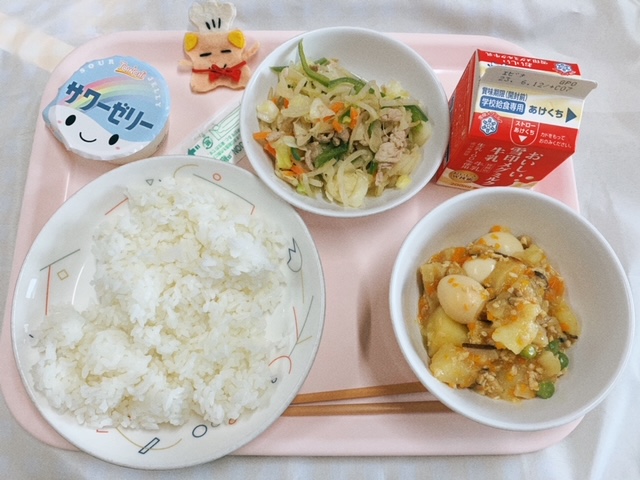 ピンクのトレーに給食が右上から反時計回りで牛乳、肉入り野菜炒め、サワーゼリー、コック帽をかぶった布製マスコット、ごはん、じゃがいものそぼろ煮と並べられている写真