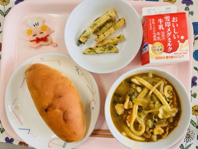 ピンクのトレーに給食が右上から反時計回りで牛乳、フライドポテト、コック帽をかぶった布製マスコット、ぶどうパン、カレーうどんと並べられている写真