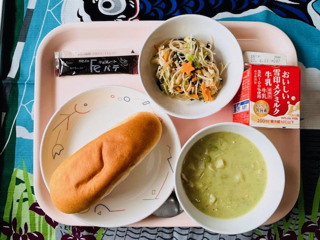 ピンクのトレーに給食が右上から反時計回りで牛乳、ビーフンソテー、チョコパテ、コック帽をかぶった布製マスコット、コッペパン、グリーンスープと並べられている写真