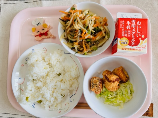 ピンクのトレーに給食が右上から反時計回りで牛乳、ごもくきんぴら、コック帽をかぶった布製マスコット、グリーンピースごはん、さけフライとキャベツソテーと並べられている写真