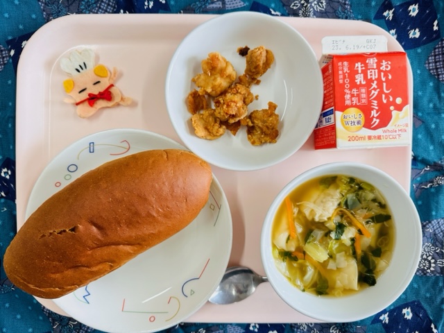 ピンクのトレーに給食が右上から反時計回りで牛乳、とりにくのうめふうみあげ、コック帽をかぶった布製マスコット、くろパン、わんたんスープと並べられている写真