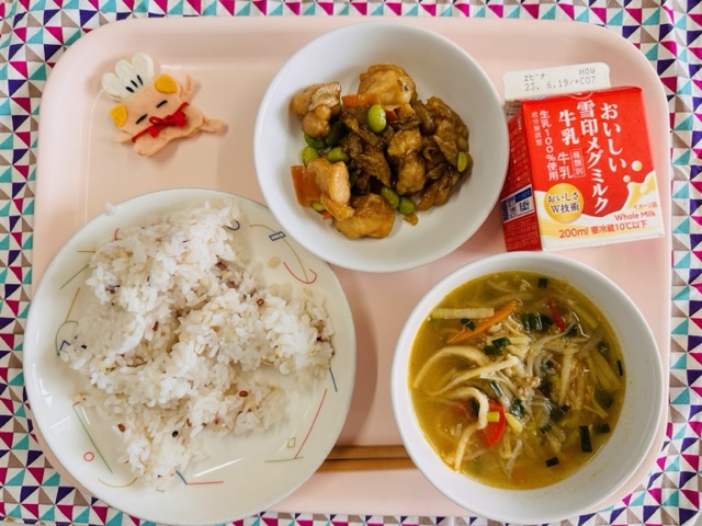 ピンクのトレーに給食が右上から反時計回りで牛乳、チキンチキンごぼう、コック帽をかぶった布製マスコット、じゅっこくまいいりごはん、きりぼしだいこんのスープと並べられている写真