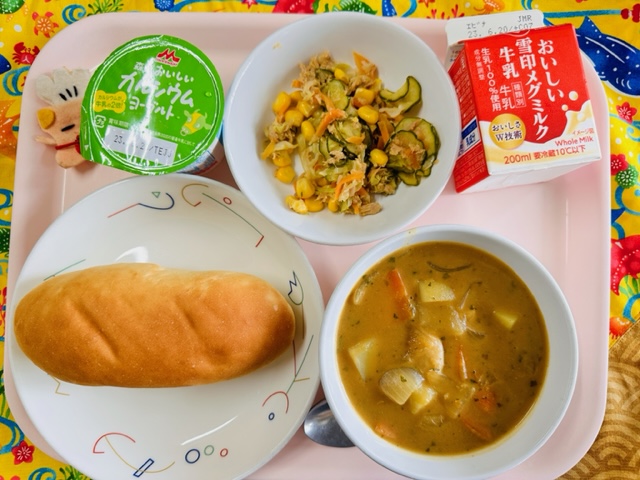 ピンクのトレーに給食が右上から反時計回りで牛乳、ツナコーンサラダ、ヨーグルト、コック帽をかぶった布製マスコット、コッペパン、カレーシチューと並べられている写真