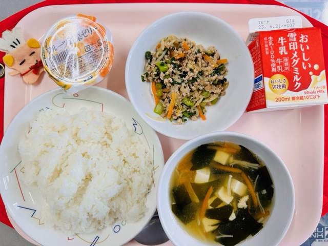 ピンクのトレーに給食が右上から反時計回りで牛乳、ビビンバの具、フルーツゼリー、コック帽をかぶった布製マスコット、ごはん、わかめスープと並べられている写真