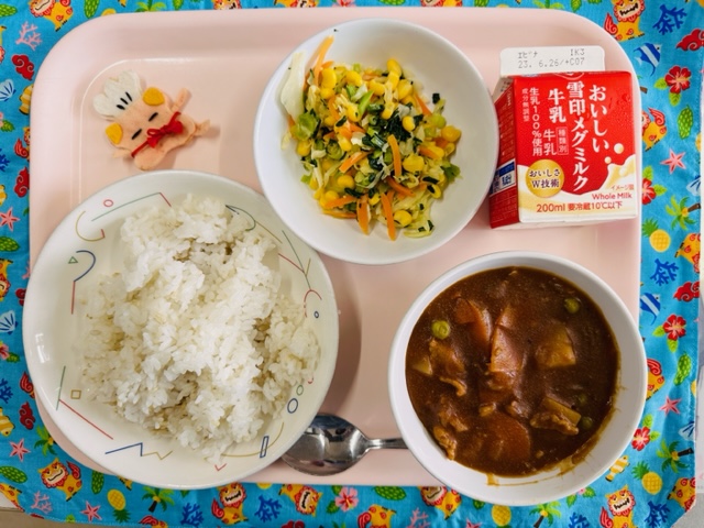 ピンクのトレーに給食が右上から反時計回りで牛乳、やさいソテー、コック帽をかぶった布製マスコット、ごはん、ハヤシライスと並べられている写真