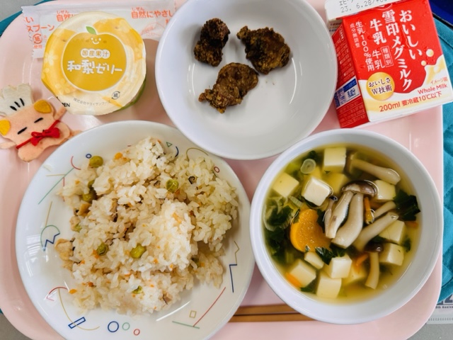 ピンクのトレーに給食が右上から反時計回りで牛乳、ぶたにくのゆかりあげ、和梨ゼリー、コック帽をかぶった布製マスコット、かしわずし、すましじると並べられている写真