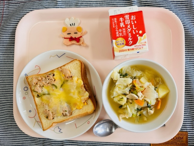 ピンクのトレーに給食が右上から反時計回りで牛乳、コック帽をかぶった布製マスコット、ツナピザトースト、やさいのスープにと並べられている写真