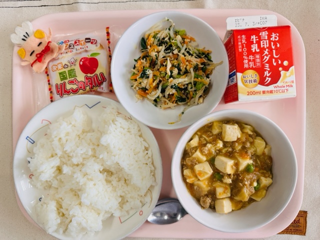 ピンクのトレーに給食が右上から反時計回りで牛乳、とりにくとやさいのナムル、国産りんごのタルト、コック帽をかぶった布製マスコット、ごはん、マーボー豆腐と並べられている写真