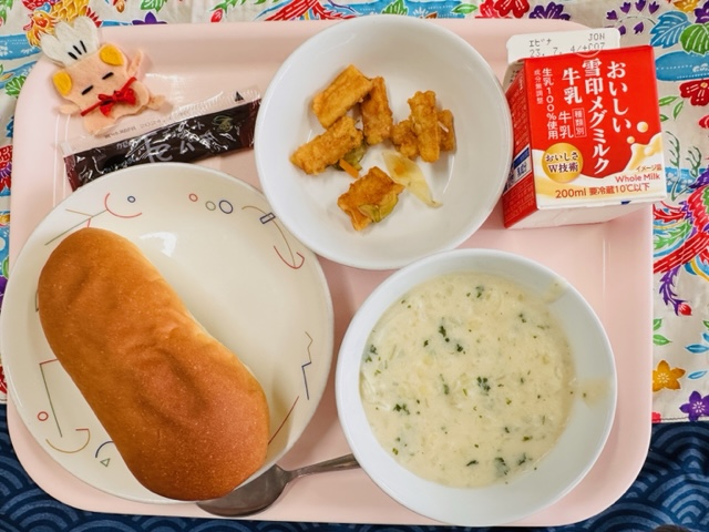 ピンクのトレーに給食が右上から反時計回りで牛乳、いかのマリネ、コック帽をかぶった布製マスコット、チョコパテ、ソフトフランスパン、ポテトスープと並べられている写真