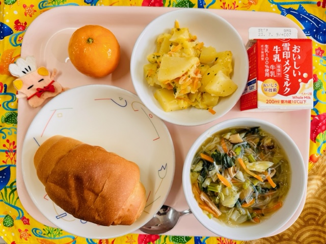 ピンクのトレーに給食が右上から反時計回りで牛乳、じゃがいものチーズやき、冷凍みかん、コック帽をかぶった布製マスコット、ロールパン、とりだんごとキャベツのスープと並べられている写真