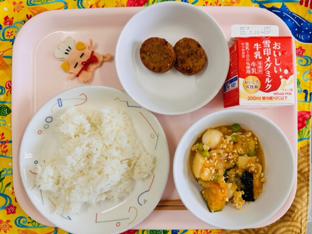 ピンクのトレーに給食が右上から反時計回りで牛乳、しろごまつくね、コック帽をかぶった布製マスコット、ごはん、かぼちゃのそぼろにと並べられている写真