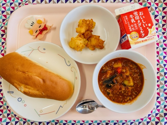 ピンクのトレーに給食が右上から反時計回りで牛乳、じゃがじゃが、コック帽をかぶった布製マスコット、ミルクパン、こくまろラタトゥイユと並べられている写真