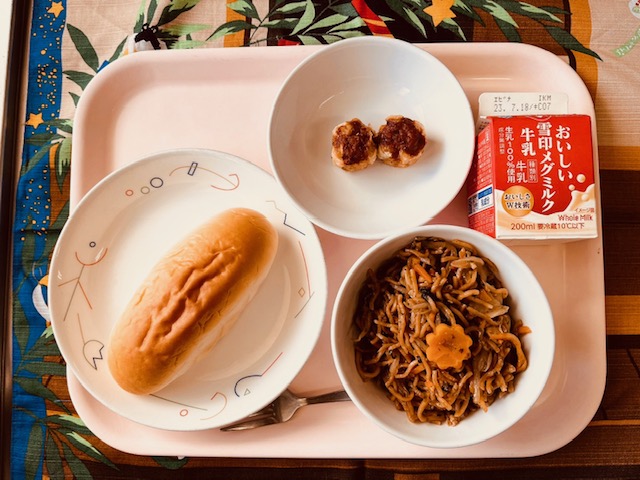 ピンクのトレーに給食が右上から反時計回りで牛乳、あげシューマイ、あじつけパン、ソース焼きそばと並べられている写真