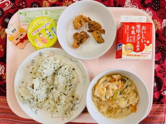 ピンクのトレーに給食が右上から反時計回りで牛乳、とりにくのハーブあげ、ゼリー、コック帽をかぶった布製マスコット、わかめごはん、たなばたじると並べられている写真