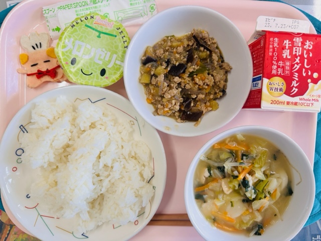 ピンクのトレーに給食が右上から反時計回りで牛乳、にくみそどんの具、メロンゼリー、コック帽をかぶった布製マスコット、ごはん、トックスープと並べられている写真