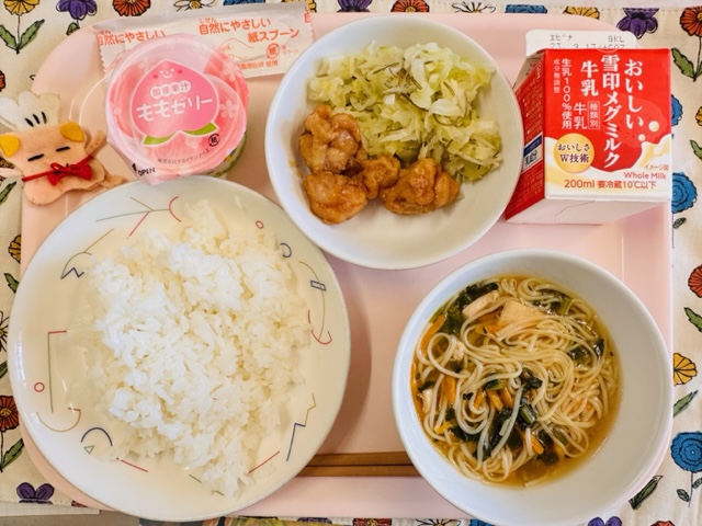 ピンクのトレーに給食が右上から反時計回りで牛乳、てりやきチキン、ももゼリー、コック帽をかぶった布製マスコット、ごはん、そうめんじると並べられている写真