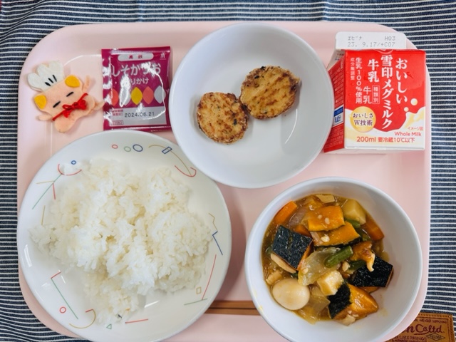 ピンクのトレーに給食が右上から反時計回りで牛乳、とりにくとやさいのフリット、しそかつおふりかけ、コック帽をかぶった布製マスコット、ごはん、かぼちゃの中華煮と並べられている写真