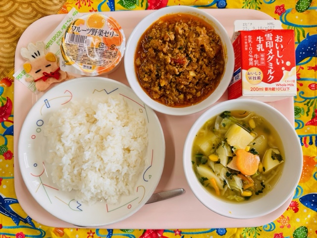 ピンクのトレーに給食が右上から反時計回りで牛乳、トマトとひき肉のアジアン、フルーツ野菜ゼリー、コック帽をかぶった布製マスコット、ごはん、やさいスープと並べられている写真