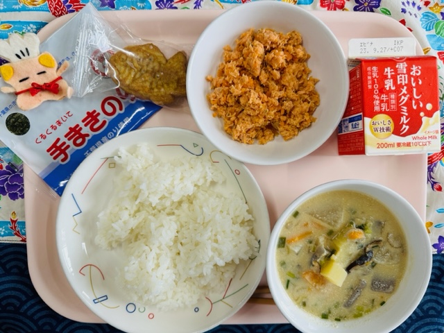 ピンクのトレーに給食が右上から反時計回りで牛乳、鮭そぼろ、ミニたい焼き、コック帽をかぶった布製マスコット、手まきのり、ごはん、ごじると並べられている写真