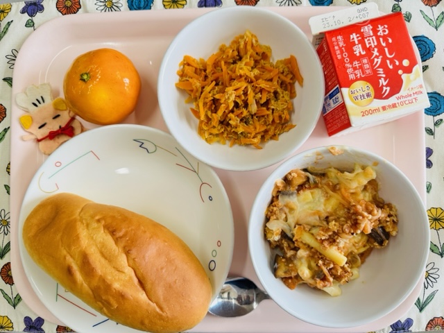 ピンクのトレーに給食が右上から反時計回りで牛乳、にんじんシリシリ、みかん、コック帽をかぶった布製マスコット、ミルクパン、ソイミートグラタンと並べられている写真