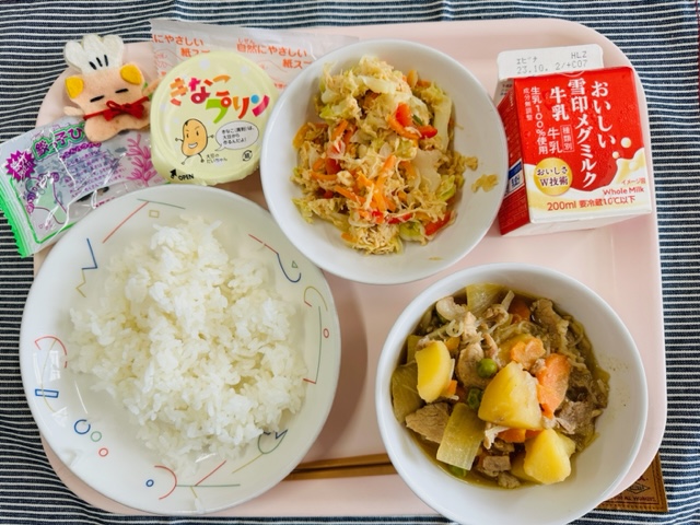 ピンクのトレーに給食が右上から反時計回りで牛乳、ささみとキャベツのサラダ、きなこプリン、コック帽をかぶった布製マスコット、ひじきのつくだに、ごはん、にくじゃがと並べられている写真