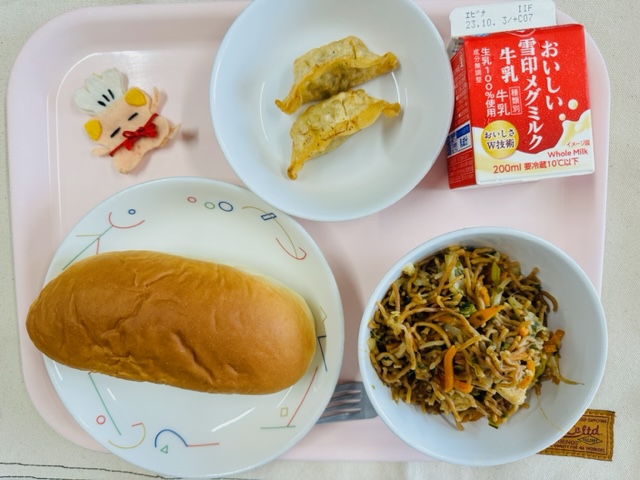 ピンクのトレーに給食が右上から反時計回りで牛乳、揚げ餃子、コック帽をかぶった布製マスコット、あじつけパン、ソースやきそばと並べられている写真